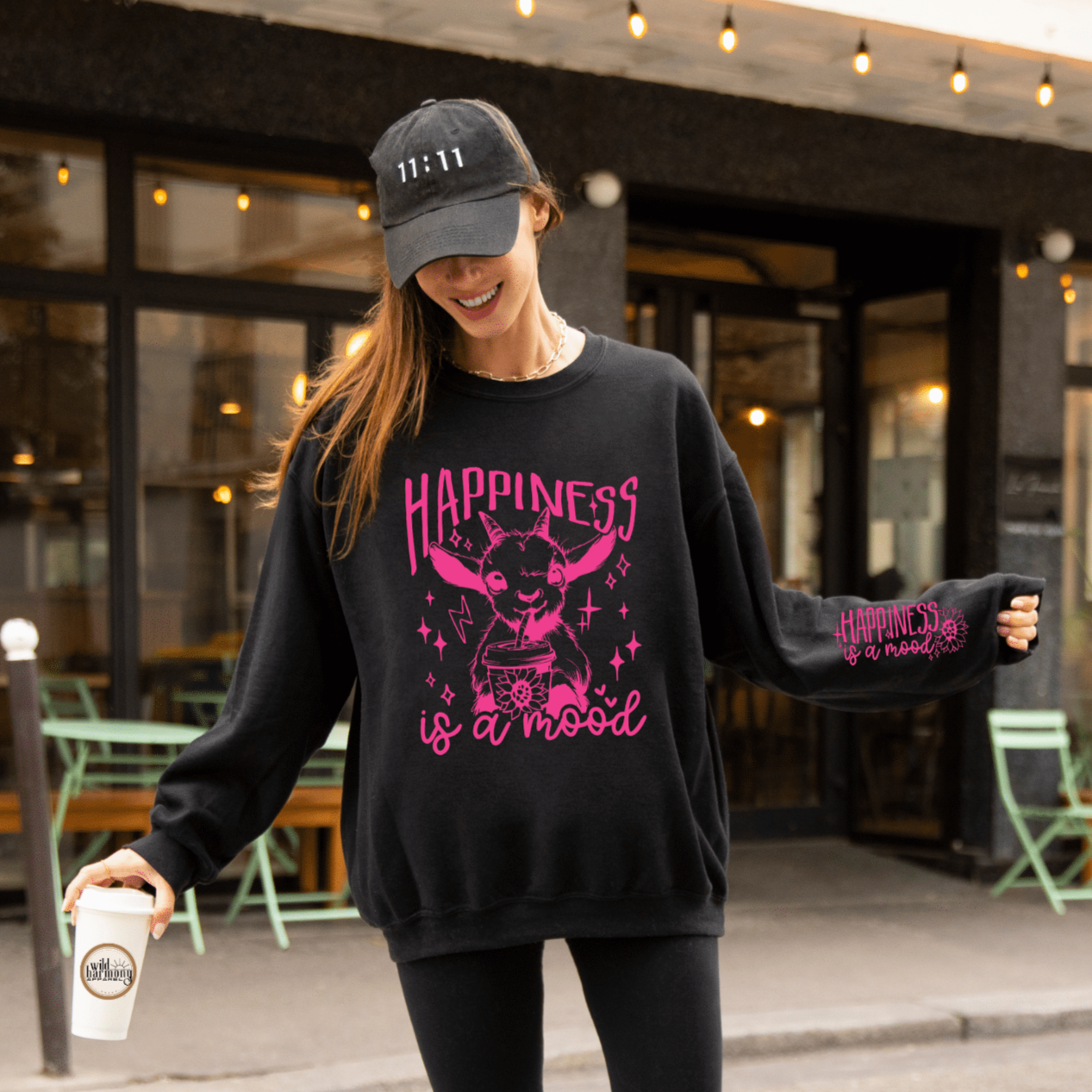 Black sweatshirt with bright pink "Happiness is a Mood" on the front with a goat drinking an iced coffee. On the left sleeve it says "Happiness is a mood"