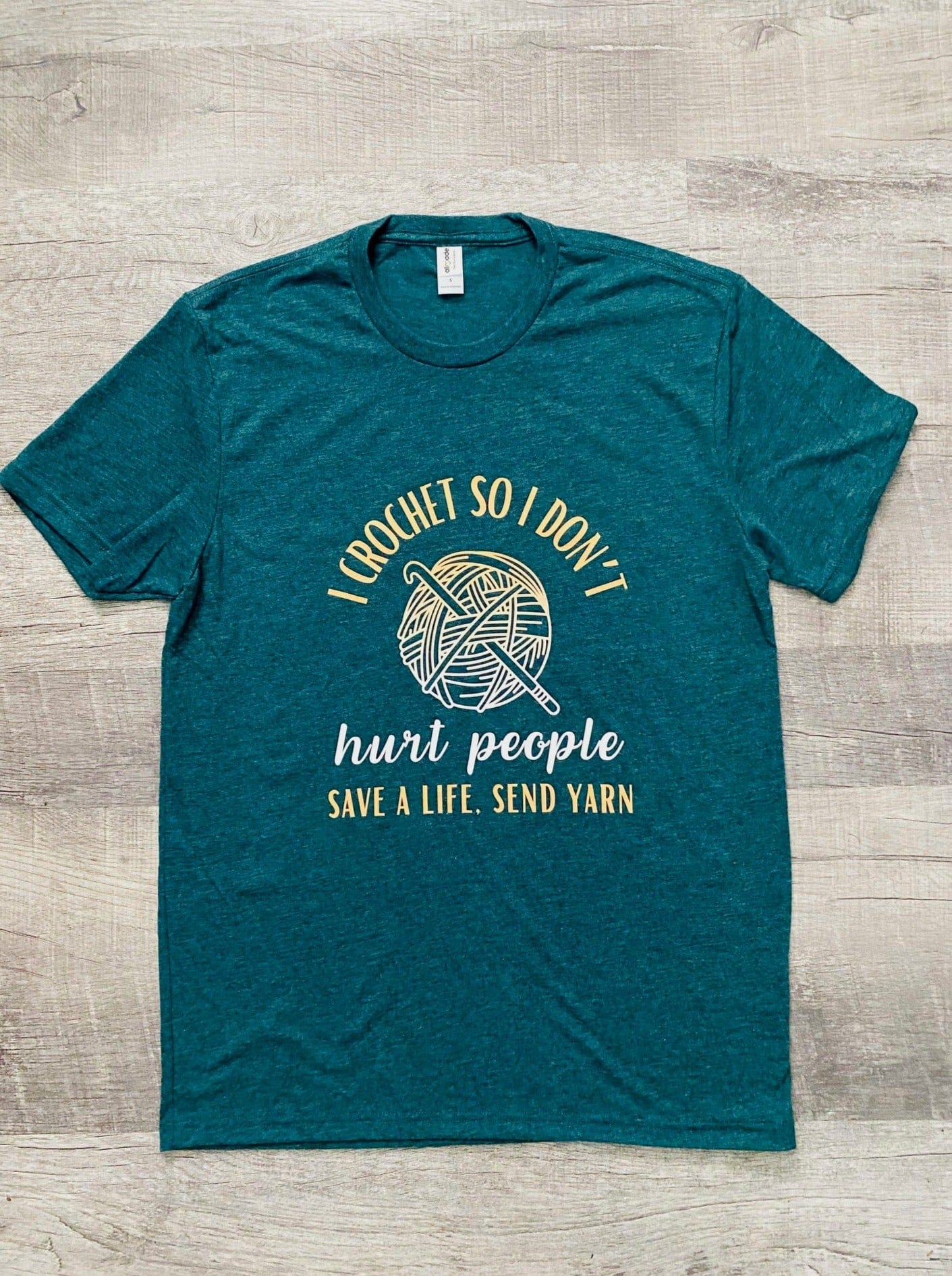 Sea Green short sleeve t-shirt with "I CROCHET SO I DON'T'" at the top in yellow and then a ball of yarn with a crochet hook in the middle that is half yellow and half white. Then below the yarn ball it says "hurt people" in white. At the very bottom it says "SAVE A LIFE, SEND YARN" in yellow.