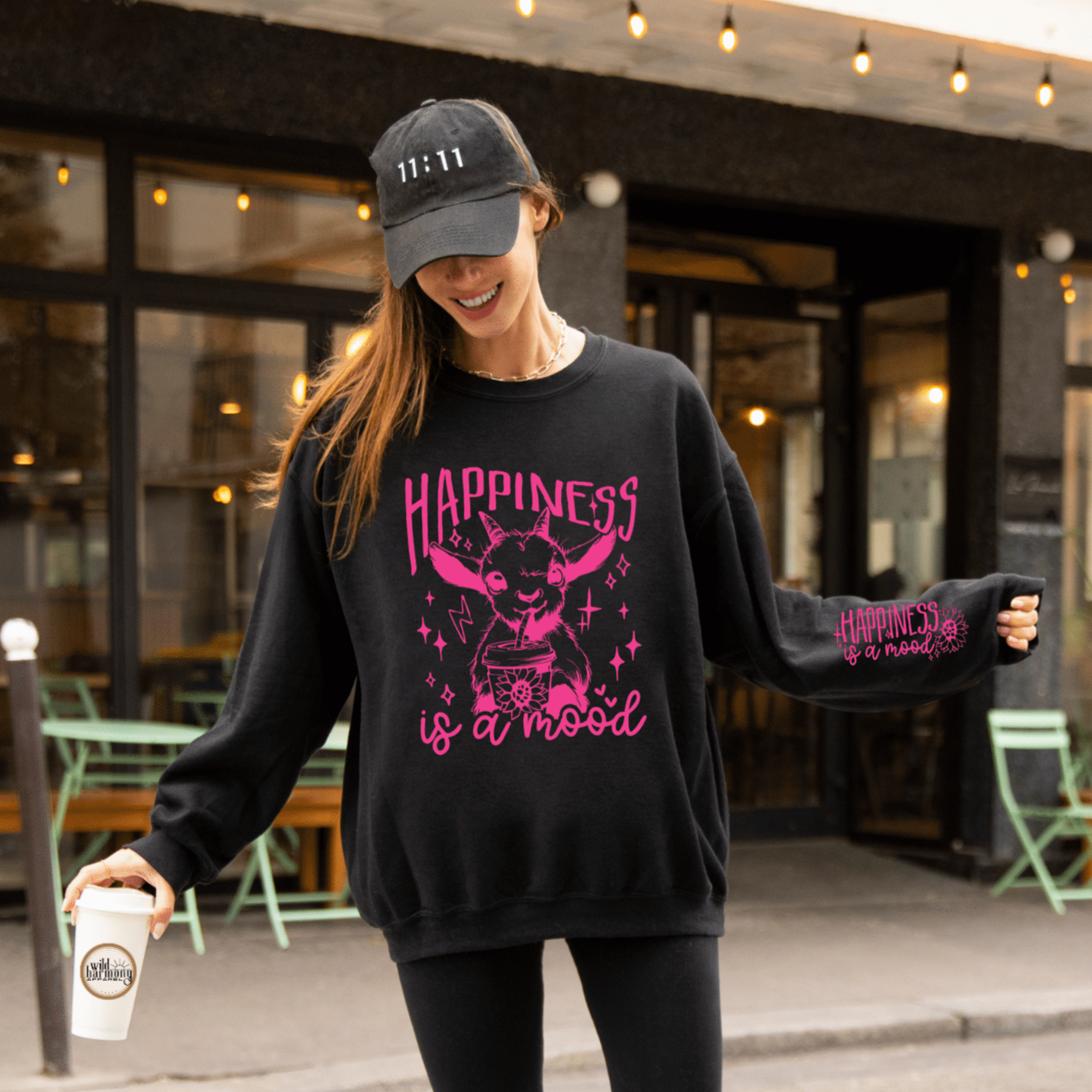 Black sweatshirt with bright pink "Happiness is a Mood" on the front with a goat drinking  an iced coffee. On the left sleeve it says "Happiness is a mood"