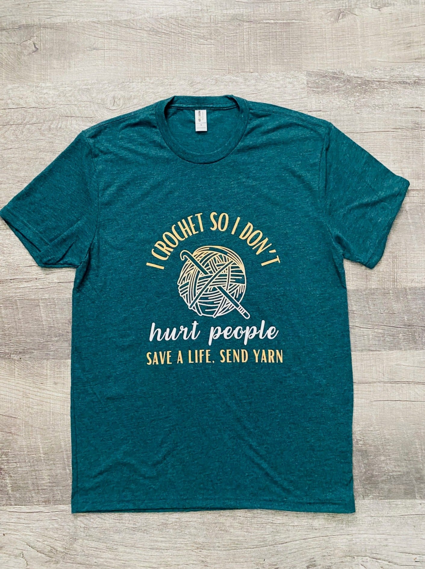 Sea Green short sleeve t-shirt with "I CROCHET SO I DON'T'" at the top in yellow and then a ball of yarn with a crochet hook in the middle that is half yellow and half white. Then below the yarn ball it says "hurt people" in white. At the very bottom it says "SAVE A LIFE, SEND YARN" in yellow. 