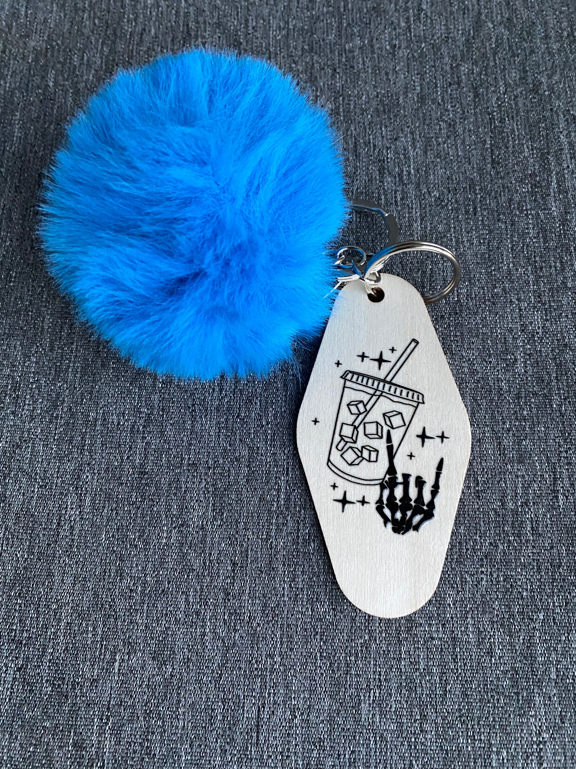 Wood keychain with an iced coffee and skeleton hand on the front. Attached to the keychain is a fuzzy ball.