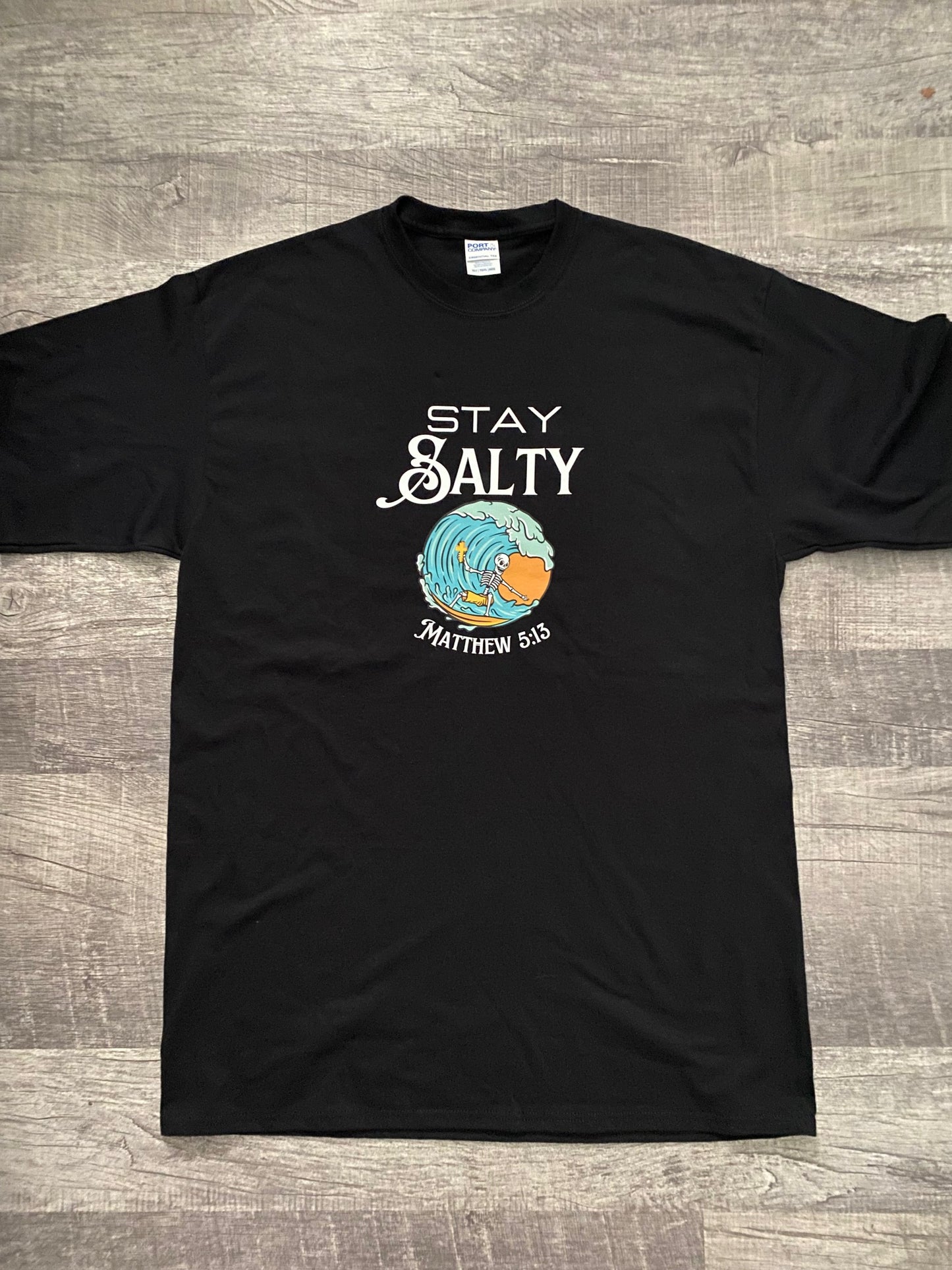 Black t-shirt with "Stay Salty" printed above a blue ocean wave with a skeleton riding the wave holding a cross in his hand. Then sun behind is orange. Underneath the wave graphic is printed "Matthew 5:13"