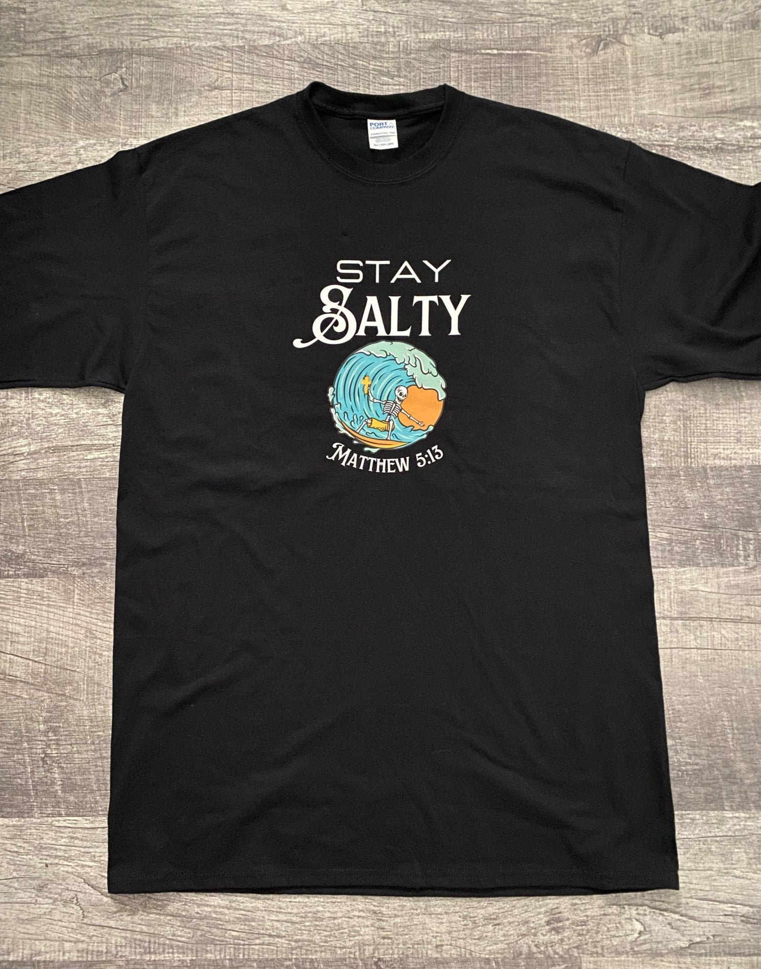 Black Tee with Stay Salty written in white above a skeleton riding a wave holding a cross. Below the wave is written Matthew 5:13. The wave is blue with orange behind it. The surfer is a skeleton wearing golden yellow shorts and riding an orange surfboard. The cross in the skeleton's hand is golden yellow. 