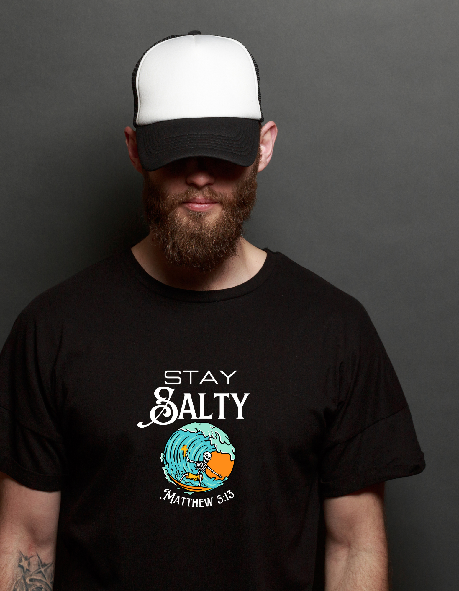Black T-Shirt with Stay Salty printed above a wave with a skeleton riding the wave on a surfboard holding a cross. Below the wave it says Matthew 5:13.