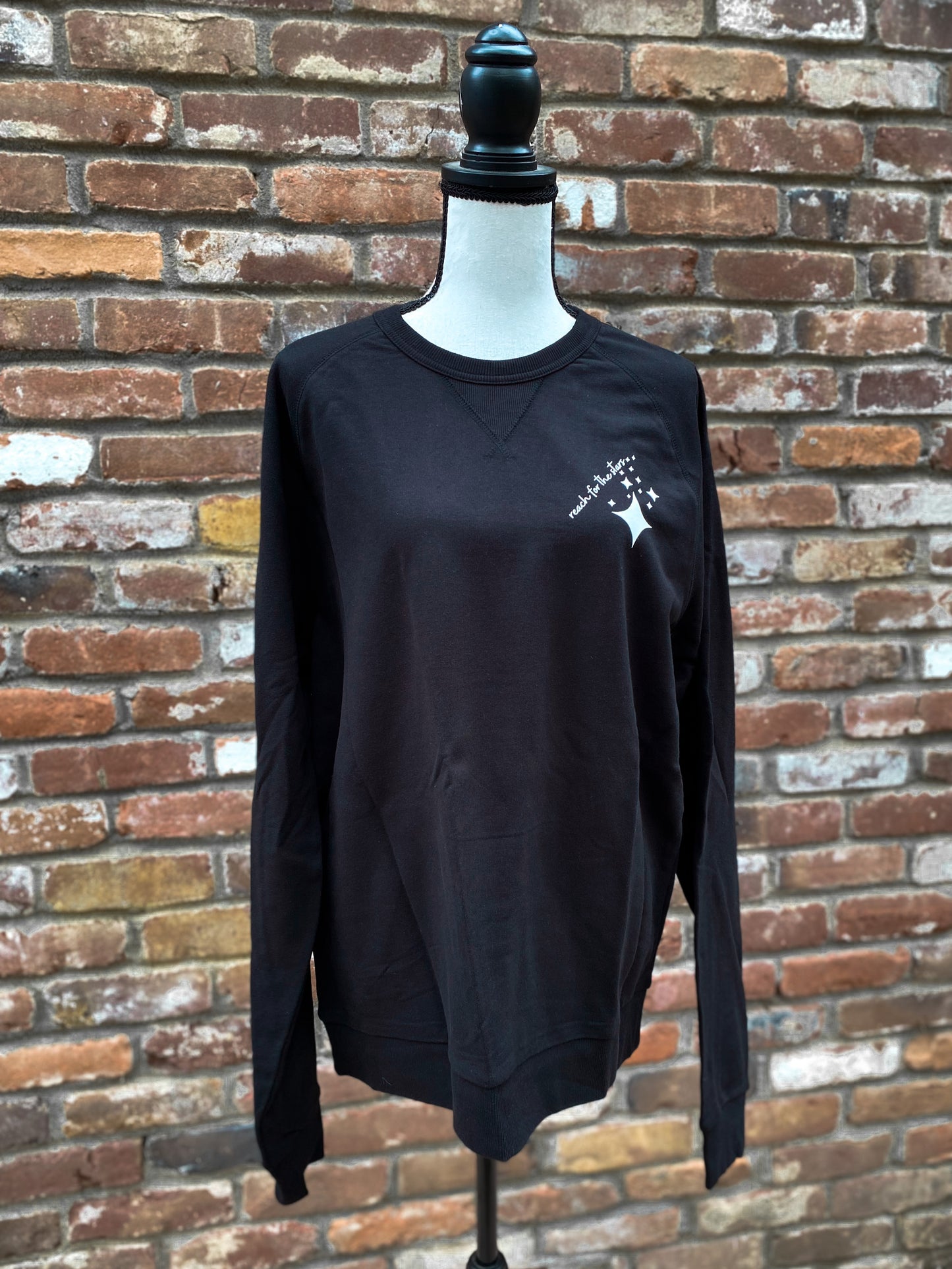 Black crewneck sweatshirt with reach for the stars written on the upper left chest with mid-century type stars underneath the written reach for the stars. Font and stars are in white and written at an angle. 